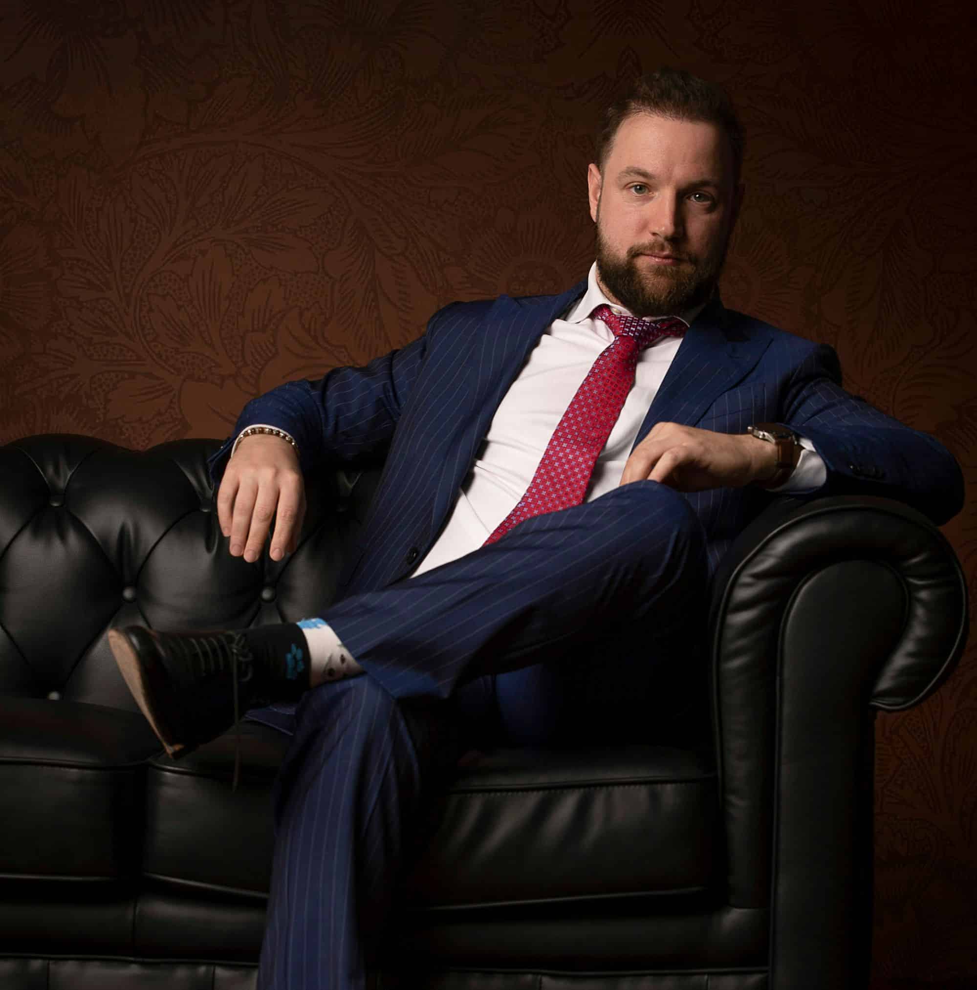 Un uomo con barba e capelli scuri, in abito blu e cravatta rossa, siede su un divano in pelle nera su uno sfondo a motivi scuri, incarnando l'etica dell'ufficio stile mentre sogna come creare un brand di moda.