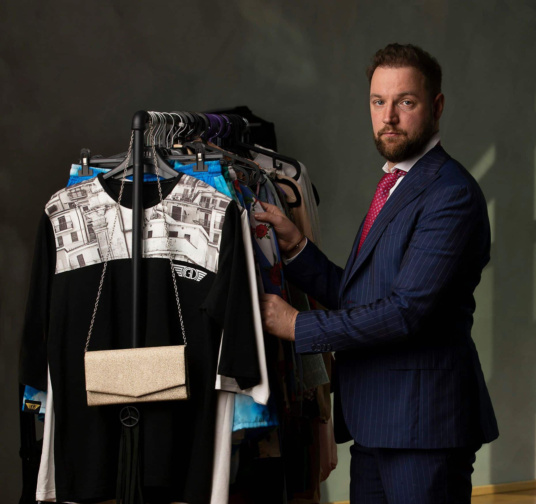 A man in a suit and tie stands next to a coat rack full of various garments, holding one of the items, as if ready to create a fashion brand.