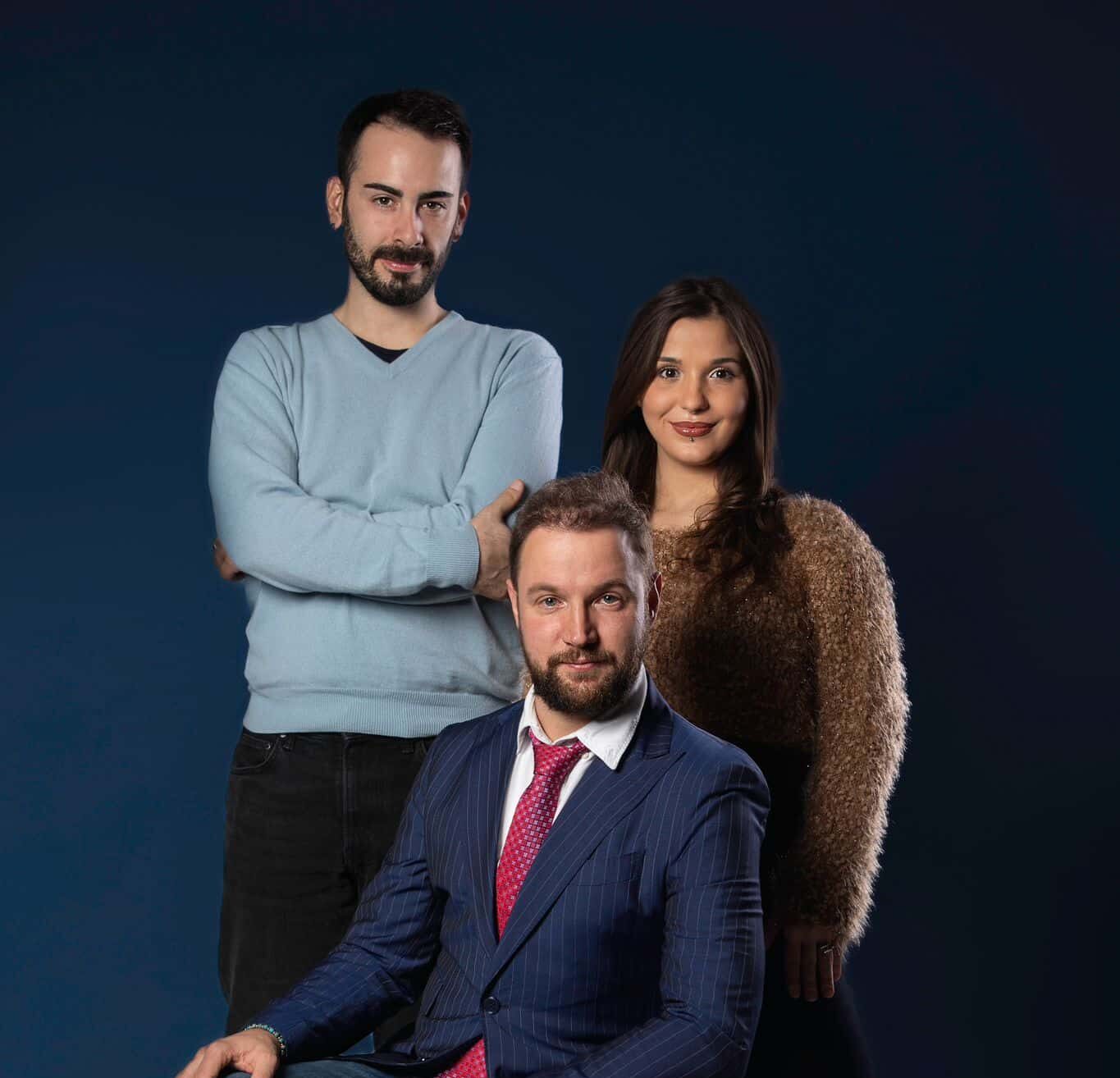 Three people pose against a dark blue background. A man in a blue jumper stands with his arms crossed, another man in a suit and jeans sits on a blue box and a woman in a furry jumper stands next to them. Their stylish outfits could inspire anyone to create a fashion brand.
