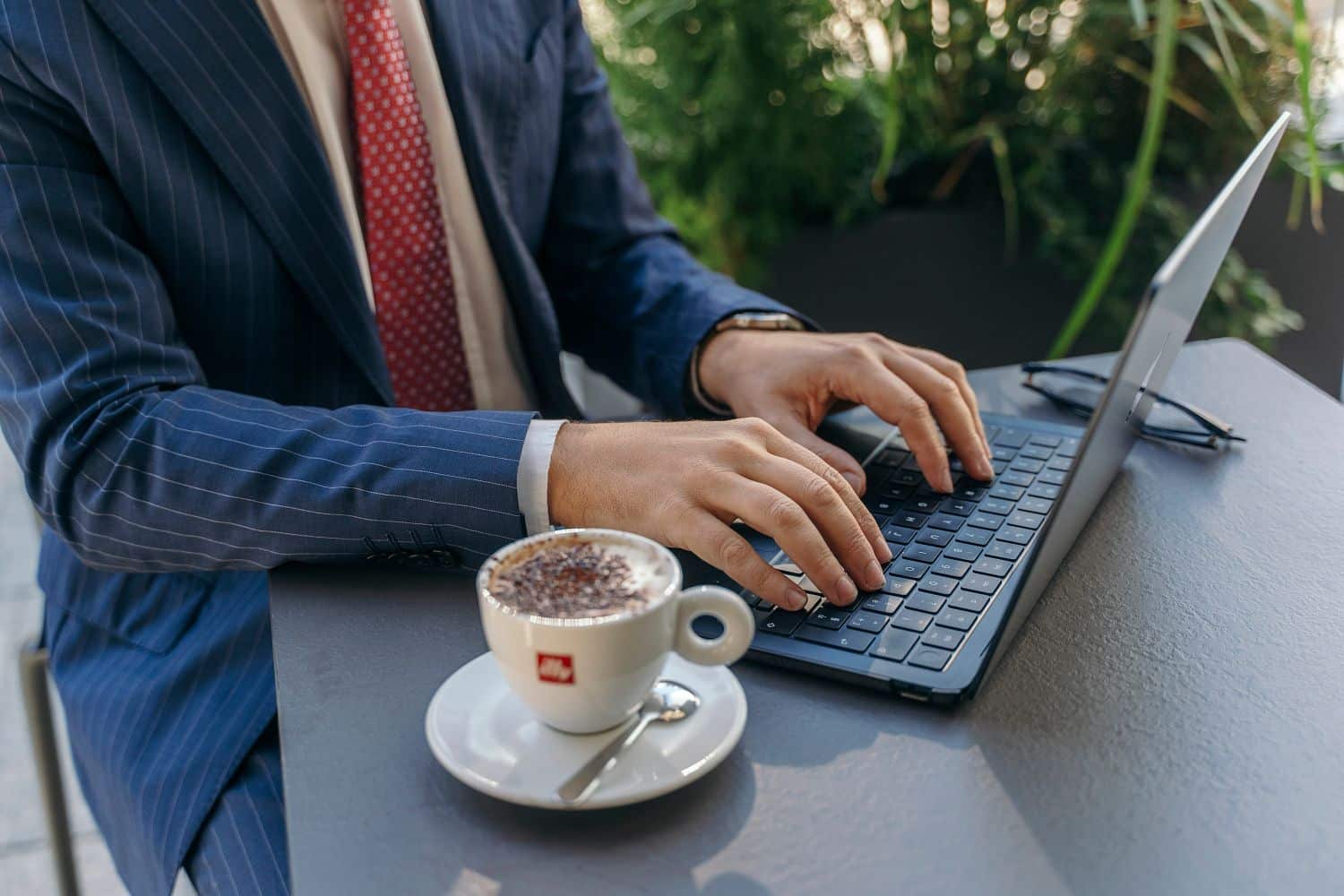 Eine Person, die auf einem Laptop tippt, sitzt an einem Tisch im Freien und hat eine Tasse Kaffee neben sich.