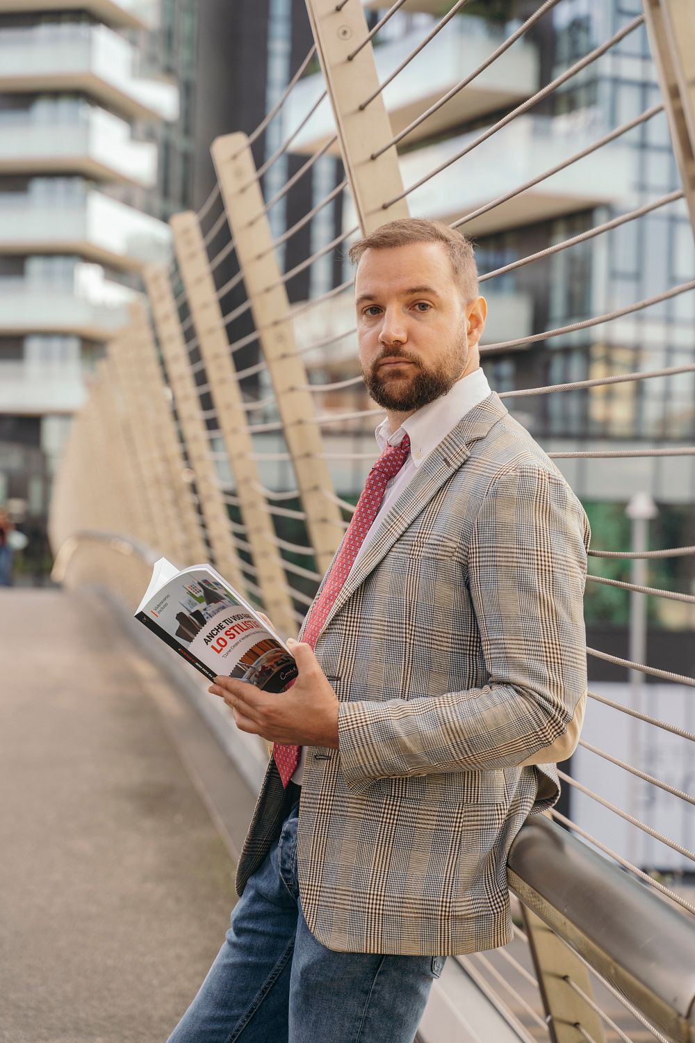 A man in a plaid jacket and red tie leans against a railing on a modern bridge, holding an open magazine. Urban buildings can be seen in the background.